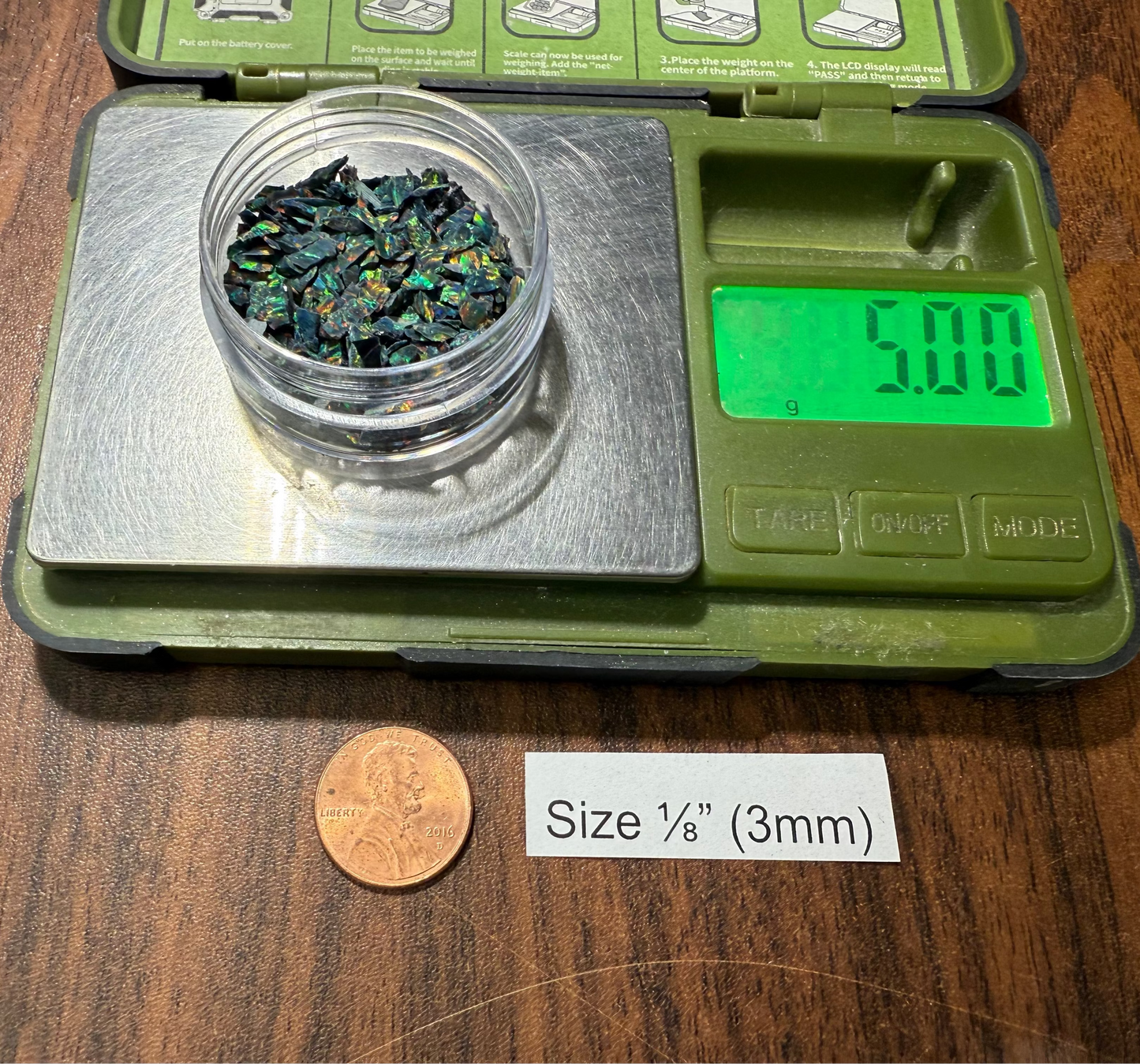 One 5g container of 1/8" opal on a digital scale with a penny for size reference on a wooden surface.